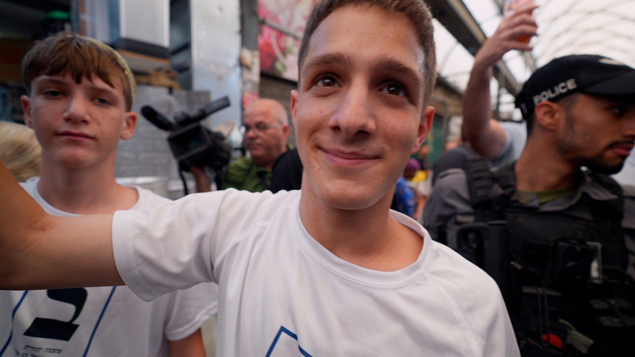 A young man in a white t-shirt stands in a busy indoors market with a police officer and a cameraman behind him, and a second young man stood to one side also in a white t-shirt