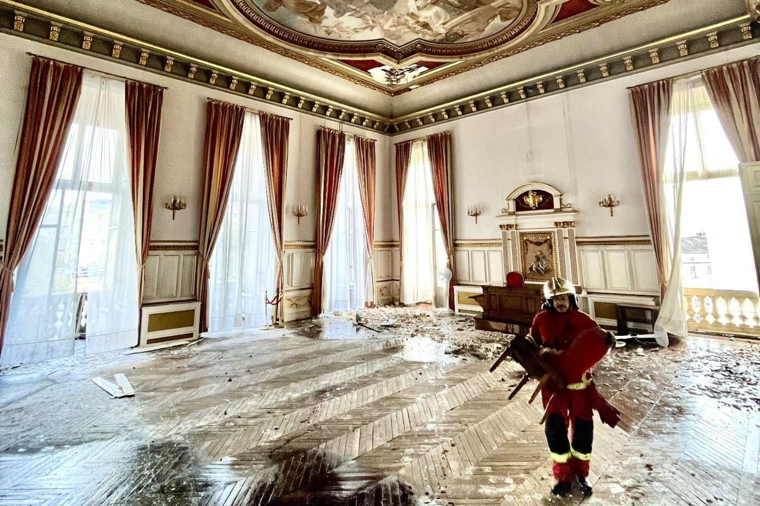 An ornate, large room in Limoges city hall. The paraquet floors are covered in rubble and a fireman is carrying a chair across the room. The ceiling has a scene of a blue sky with angels and divinities painted on it.