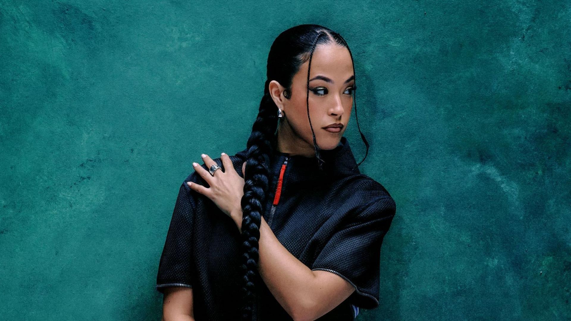 Jazzy is a 30-year-old woman with long dark hair which she wears in a braid down to her middle, with two smaller braids shaping her face. She looks over her left shoulder, her left arm across her body. She's photographed wearing a dark short sleeved shirt with a red zip against a blue backdrop.