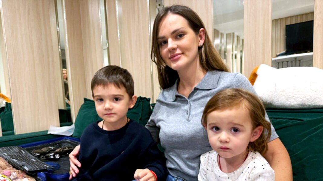 A woman with long brown hair sits on a green sofa with two young children, a boy and a girl.