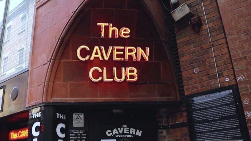 Exterior entrance to Liverpool's Cavern Club on Mathew Street, featuring the illuminated "The Cavern Club" sign above the famous arched doorway, with brick walls and information boards on either side of the entrance