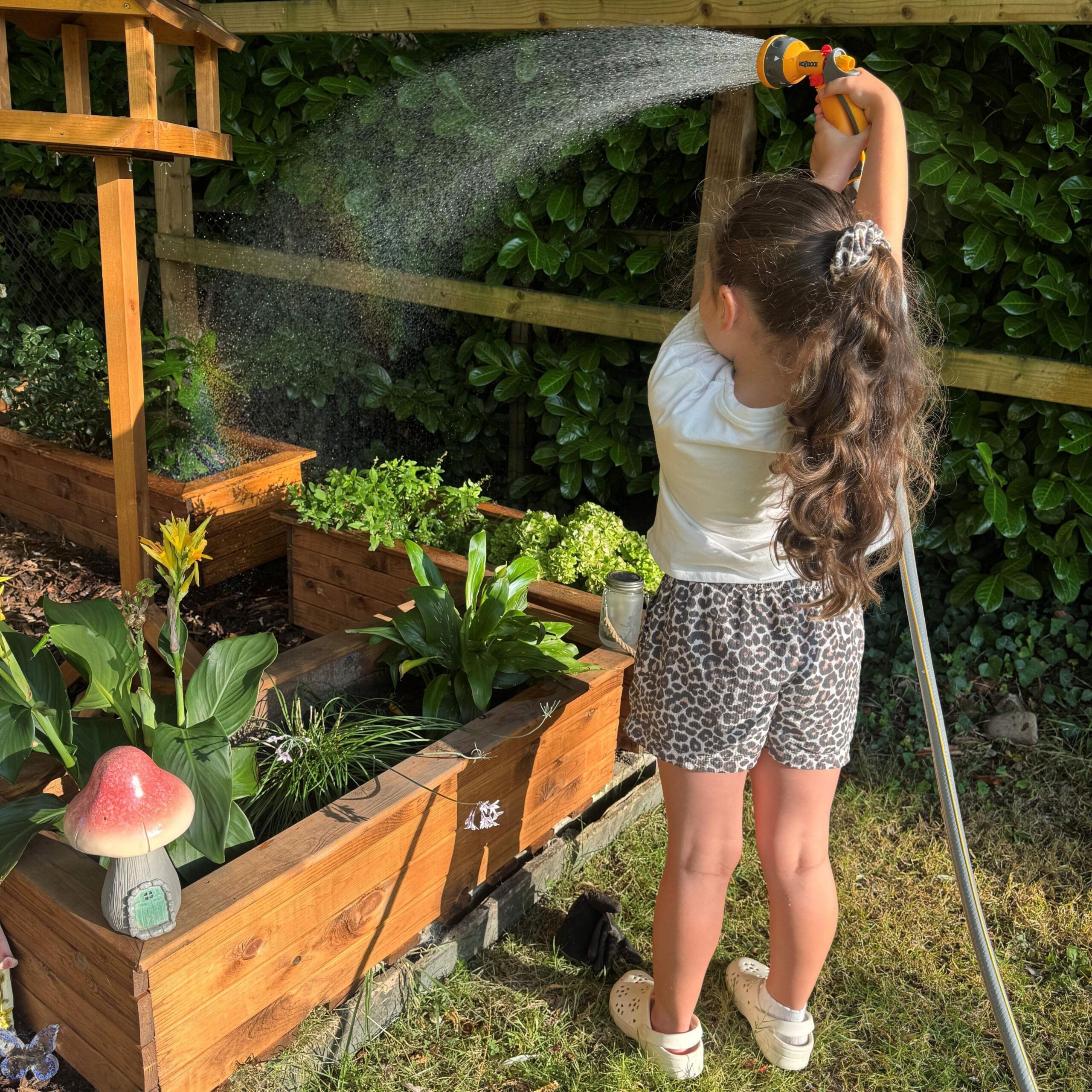 Child standing in a garden in the foreground, holding a hose spray nozzle above their head and watering raised wooden planters. Water arcs from the nozzle across the planters, which contain various flowers and leafy plants. A wooden bird-table-style structure stands beside the planters, and a mushroom-shaped garden ornament is visible at the corner of one planter. A wooden fence and dense hedging form the background.