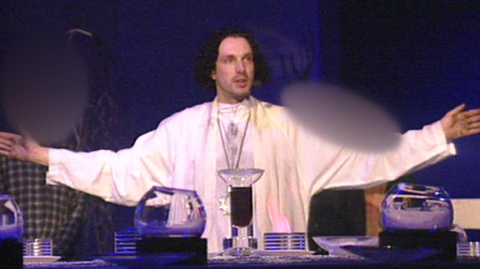 Chris Brain leads a service, wearing white robes and with his arms out wide. The lighting is dim with a blue hue.