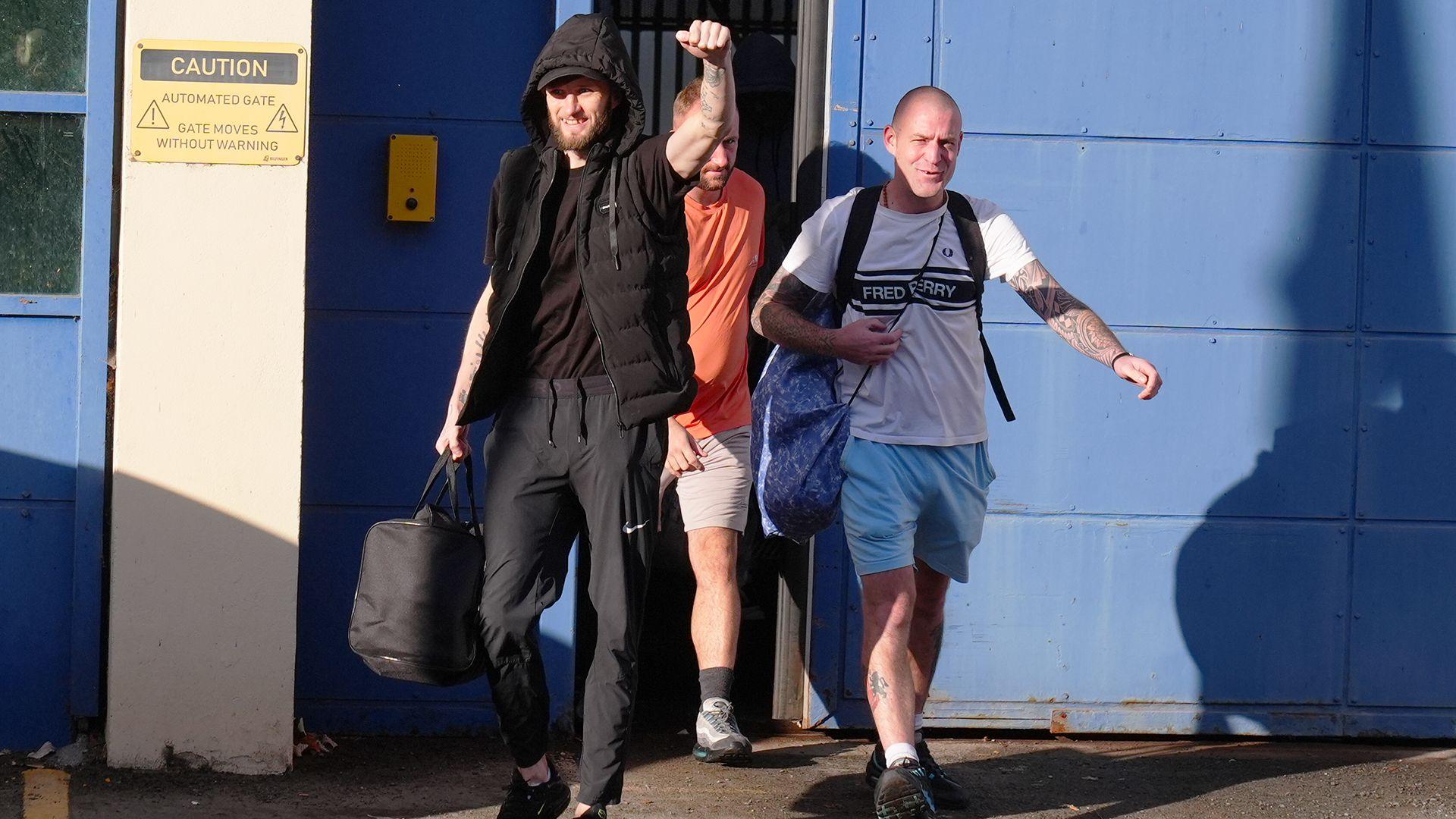 Three men leaving HMP Durham in north east England on 13 October. The group of three are in bright sunshine and smiling. The man in front is bearded and all in black with his had raised in a victory punch. Behind him is a bald-headed man wearing blue shorts and a white "Fred Perry" t-shirt and behind him is a blond man wearing stone-coloured shorts and an orange t-shirt