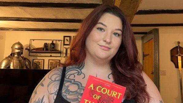 A photo of a woman with red hair smiling and holding a red book with yellow writing on the front cover. She has got tattoos on and a black tank top.