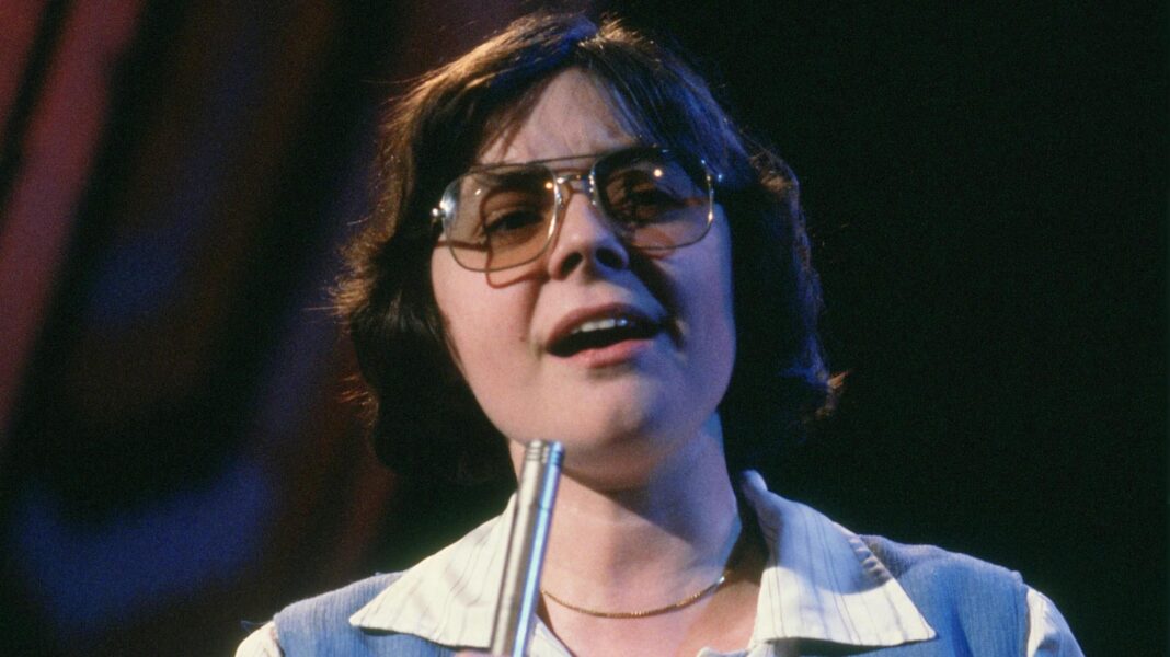 Lena Martell on Top of the Pops. She is wearing a white shirt and blue wool tank top. She has short dark hair and glasses and is singing into a microphone.