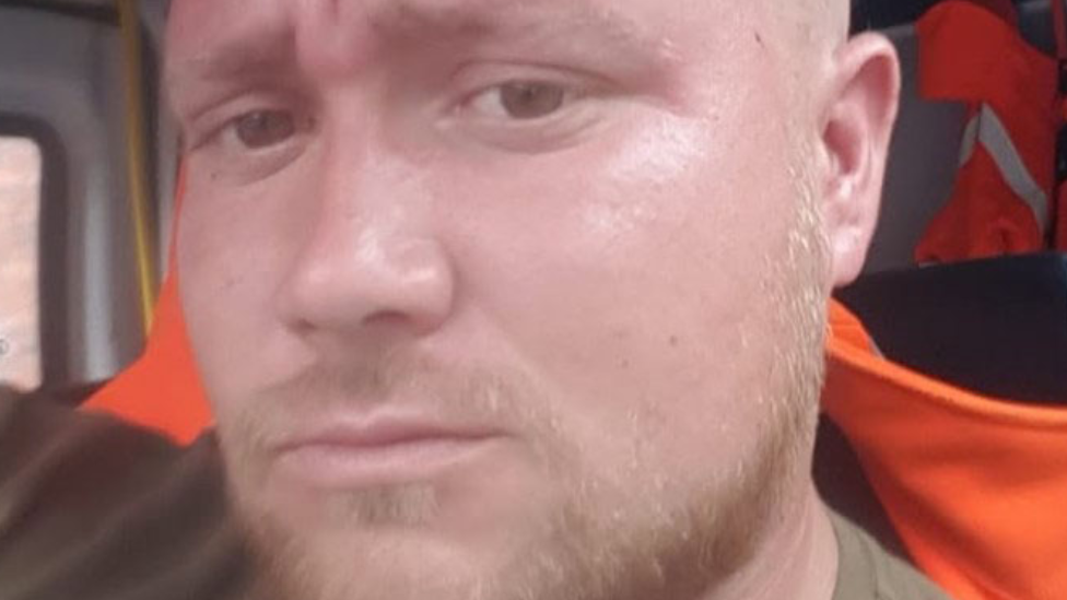 A selfie of a man with a fair beard, sat in a van with a high vis orange jacket on the seat behind him, He is looking at the camera with a serious expression.
