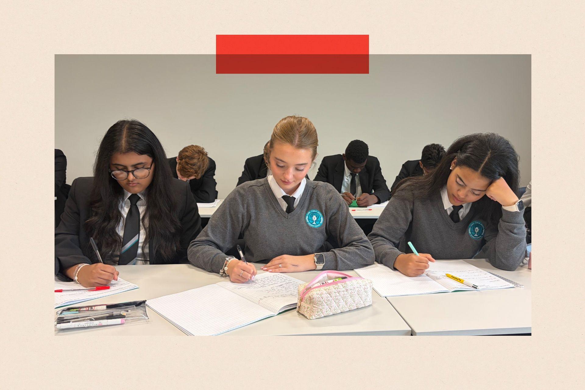 Three teenage female students sat at desks studying. The one on the left is wearing a dark blazer and the other two a grey jumper over their shirt and tie