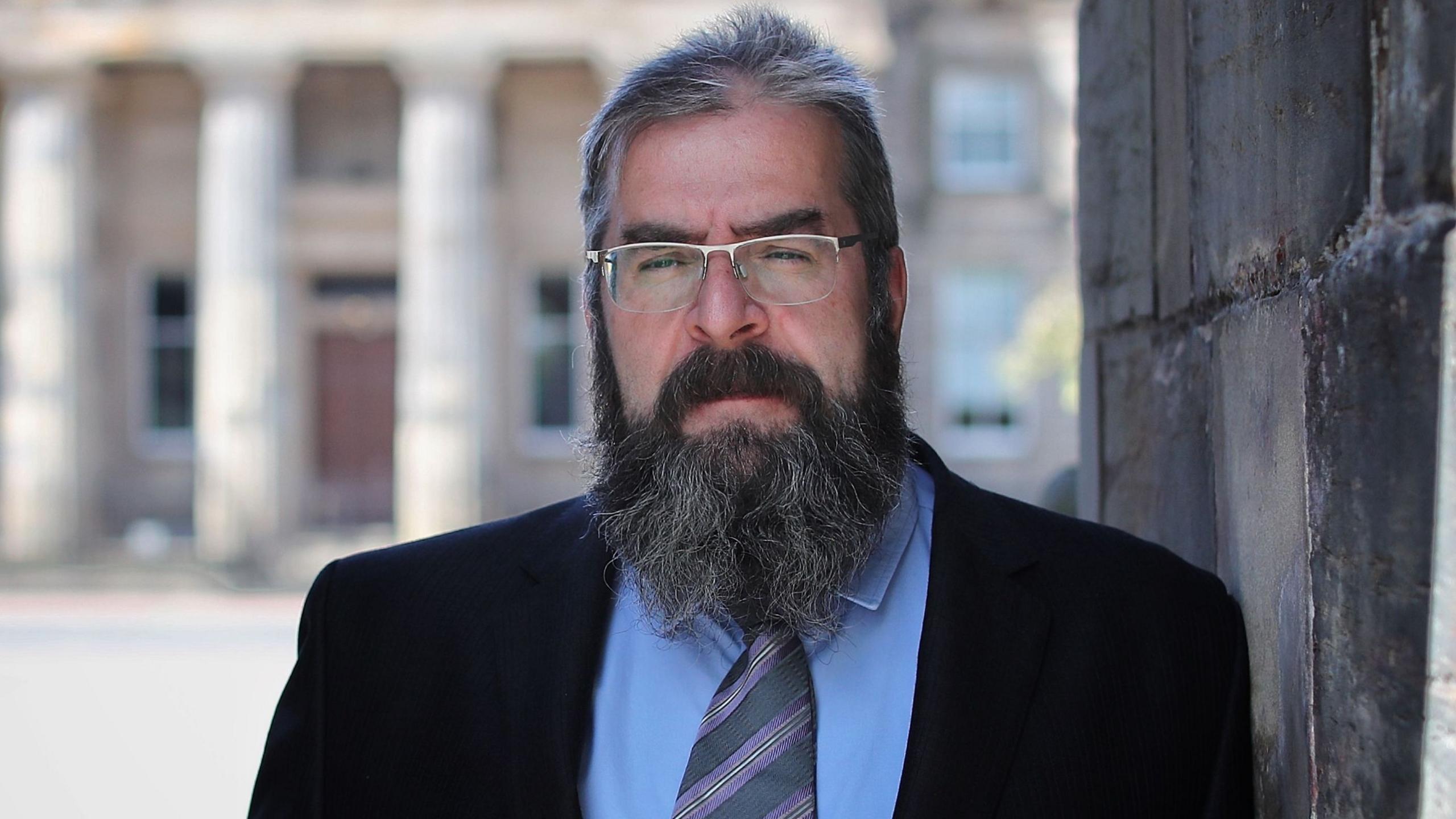 Head and shoulders shot of Tony Lenehan KC, who has short greying hair and a large busy beard. He is wearing a black suit jacket, blue shirt and purple tie.