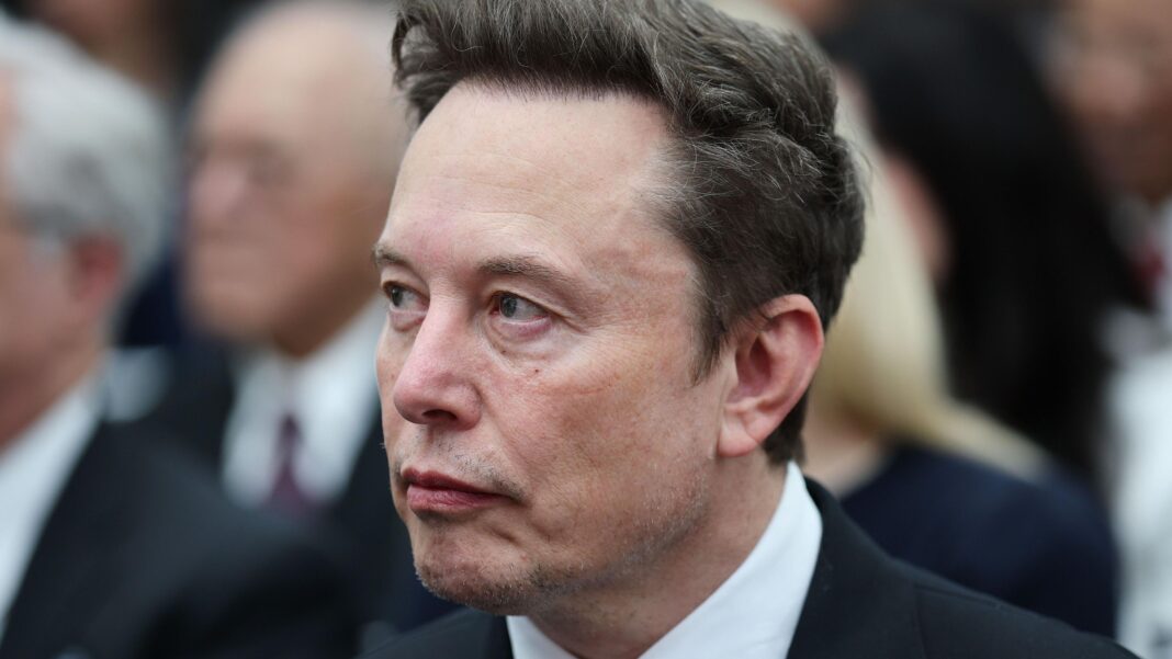 Elon Musk looks sideways as he wears a dark suit, white shirt and grey tie, with other people visible in the background.