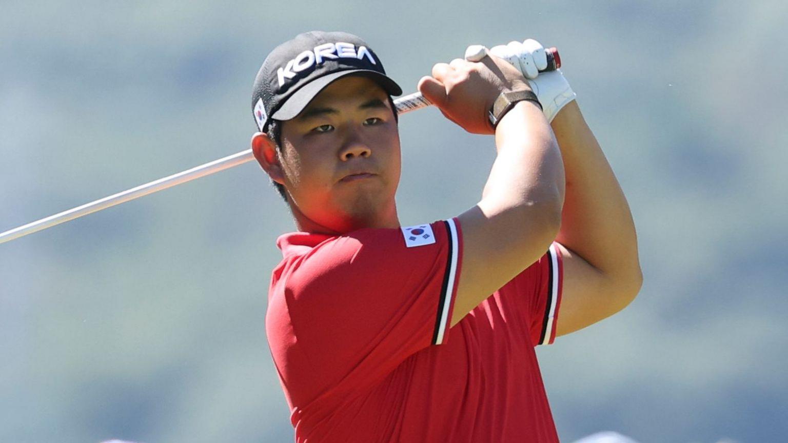 Tom Kim in a red polo shirt and a black cap, both featuring badges with the flag of South Korea. His club is behind his head at the conclusion of his swing