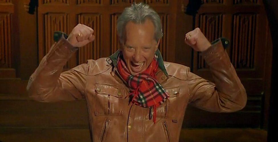 Richard E Grant raising his arms and cheering at the round table on Celebrity Traitors