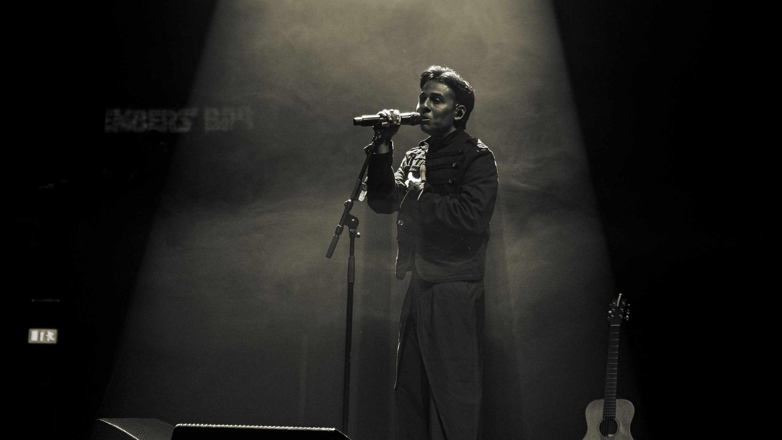 Anuv performs on stage with a spotlight over him. His guitar can be seen resting behind him and he wears a coordinated black suit outfit