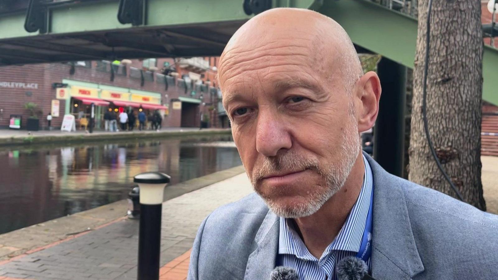 A middle aged man with a grey beard and bald head stands by a canal. He is wearing a light blue suit and striped blue shirt.