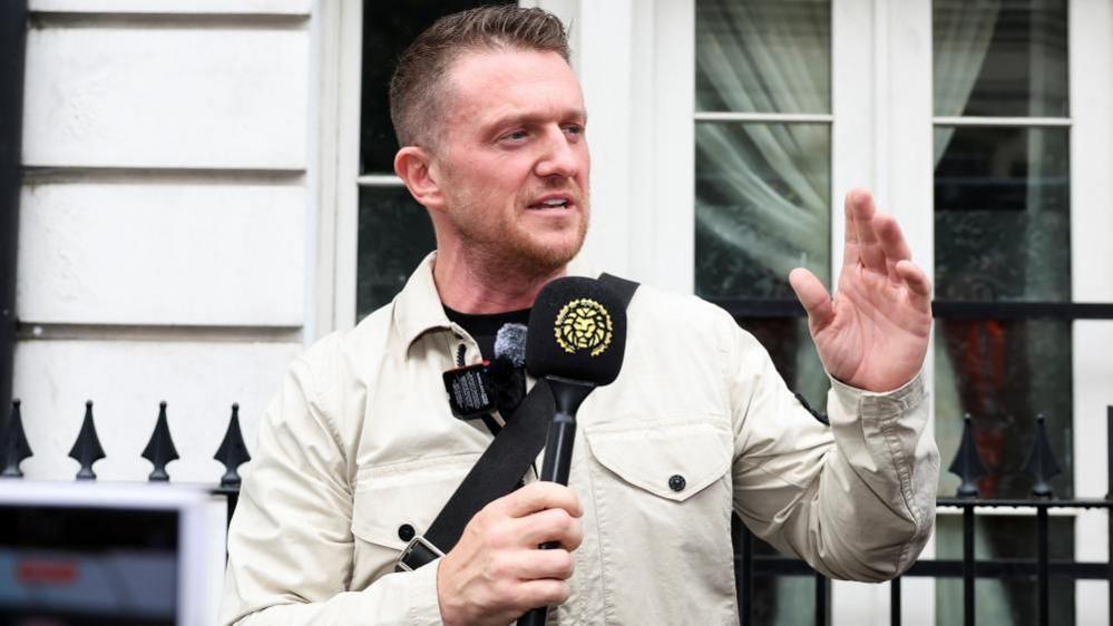 Tommy Robinson, standing outside Westminster Magistrates' Court, in London, talking into a black microphone, with a golden lion head on it, with his left hand raised. He is wearing a light jacket, a black T-shirt and has short dark hair and short beard. He is standing by black railings.
