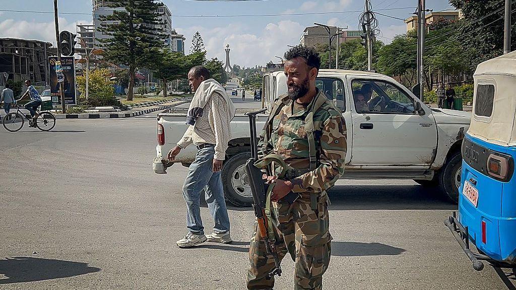 A member of the Ethiopian pro-government Tigray Peace Force (TPF) monitors activity on a street while holding a gun. He is in a camo jacket and trousers. A man wearing a white jumper and jeans walks behind him.