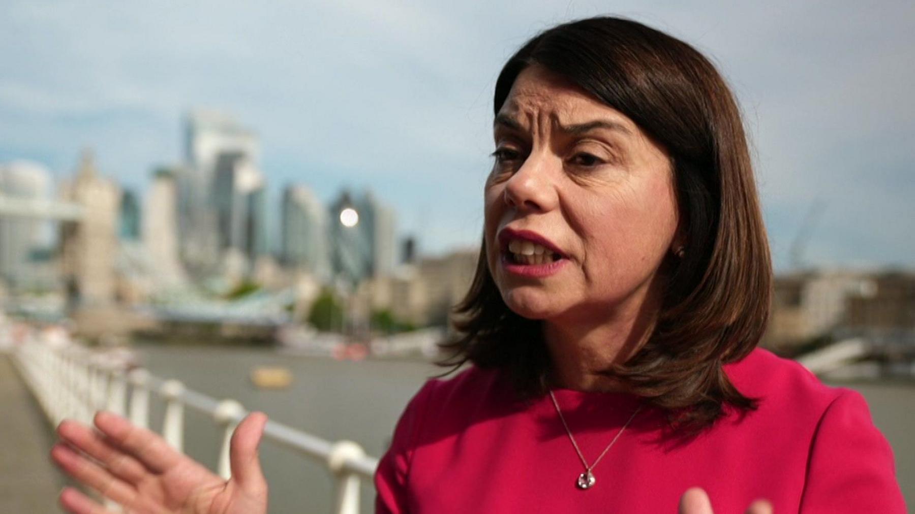 A woman in a red dress with medium length brown hair being interviewed, standing by the River Thames.