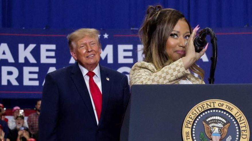 Nicki Minaj at the microphone at a podium with the presidential seal. She has a slight smile and a gold suit with a Gucci logo. Trump smiles behind her.