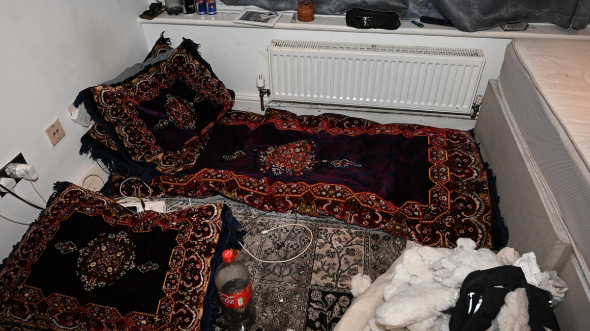 A dark coloured bed sheets and pillows are pictured lying on the floor, next to a bottle of coke. There is a mattress pictured with no bed sheets.