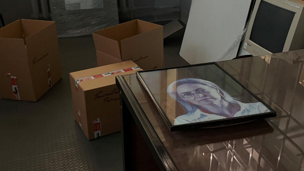 A framed photograph of Anna Politkovskaya lies on a desk in the foreground. Three cardboard boxes sit on the floor behind the desk. An old-fashioned computer monitor can also be seen in the top right hand corner of the picture.