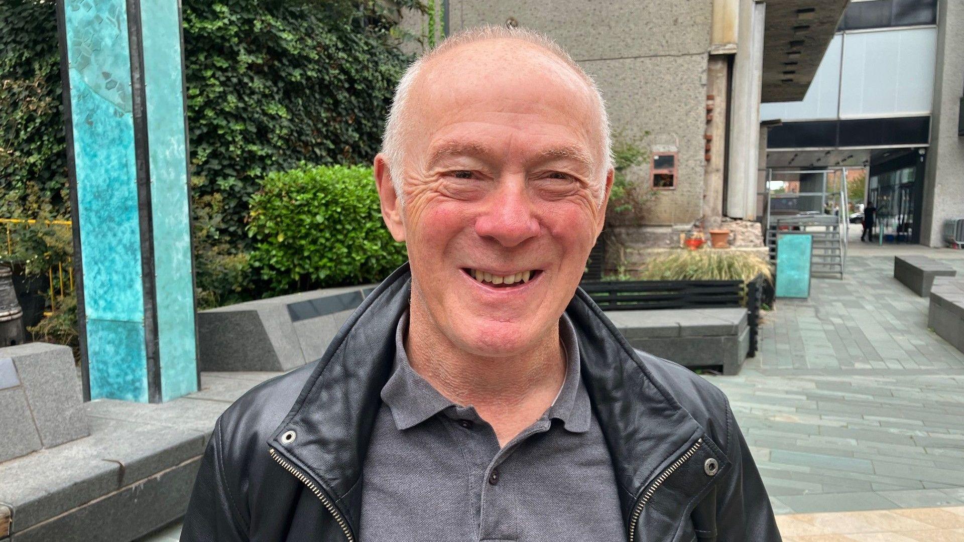 Richard Leese stands in front of stone buildings outside on a high street. He wears a grey polo shirt and black leather jacket. He has short grey hair and is smiling.