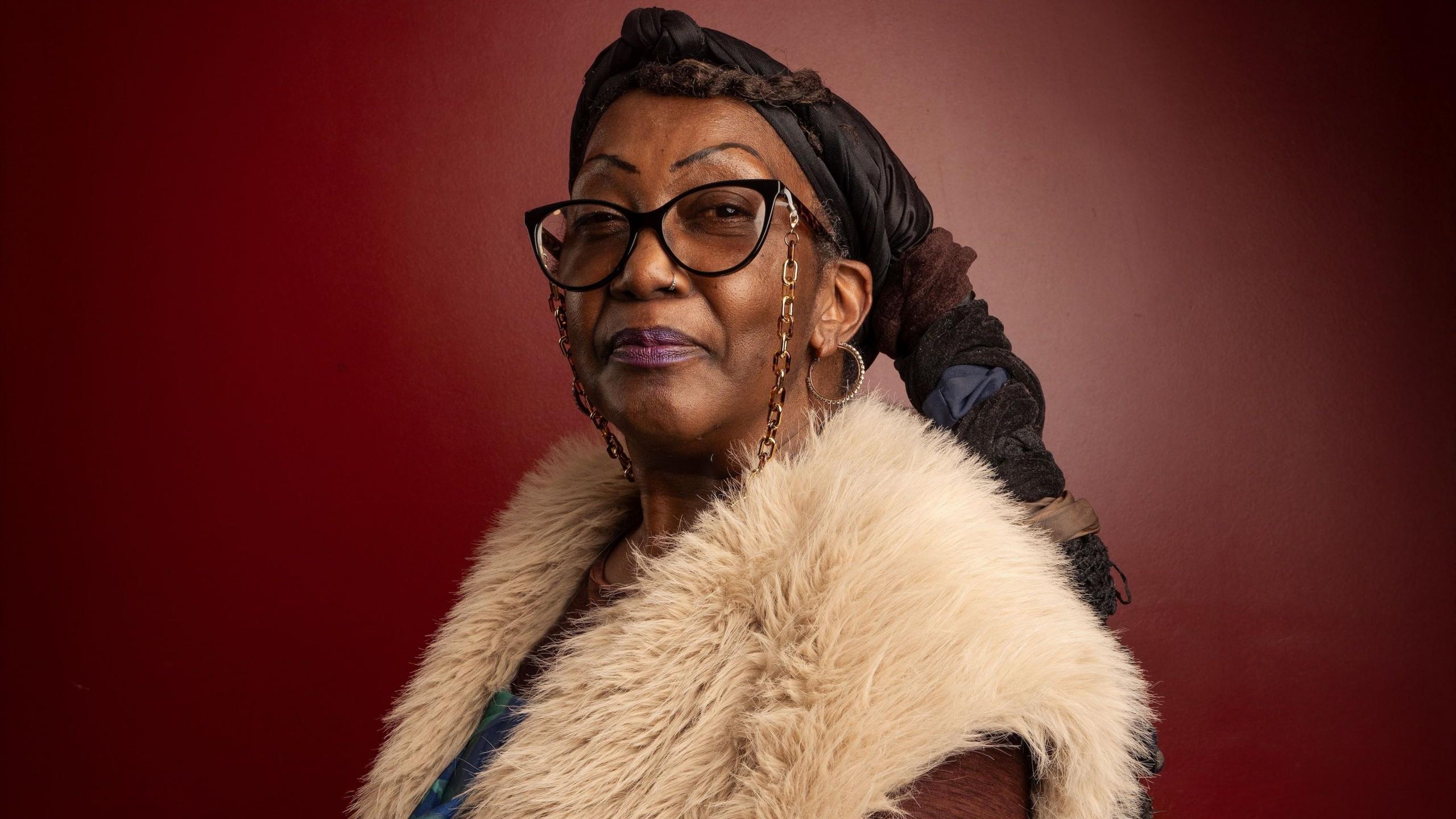 A woman standing against a deep red background, wearing glasses, a black headscarf, a cream faux-fur gilet and multiple bracelets. Long braided hair extends down the back, and one hand is held forward toward the camera.