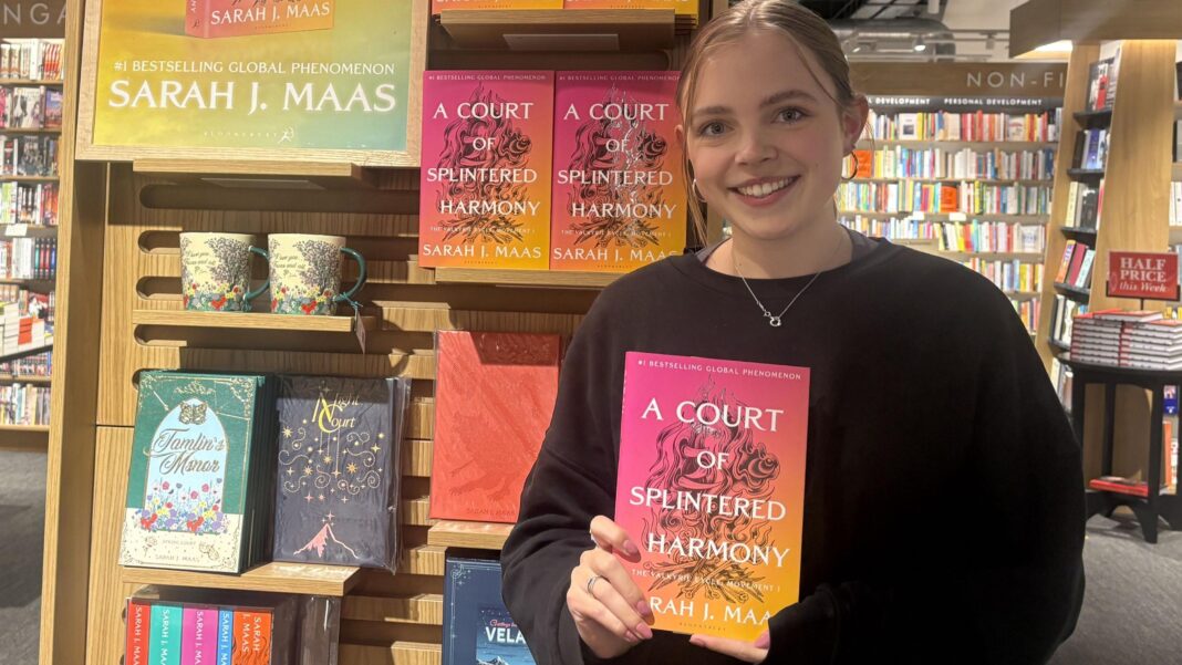 A young woman wearing a black jumper and her hair tied back is smiling, holding a colourful book in front of a bookshop display