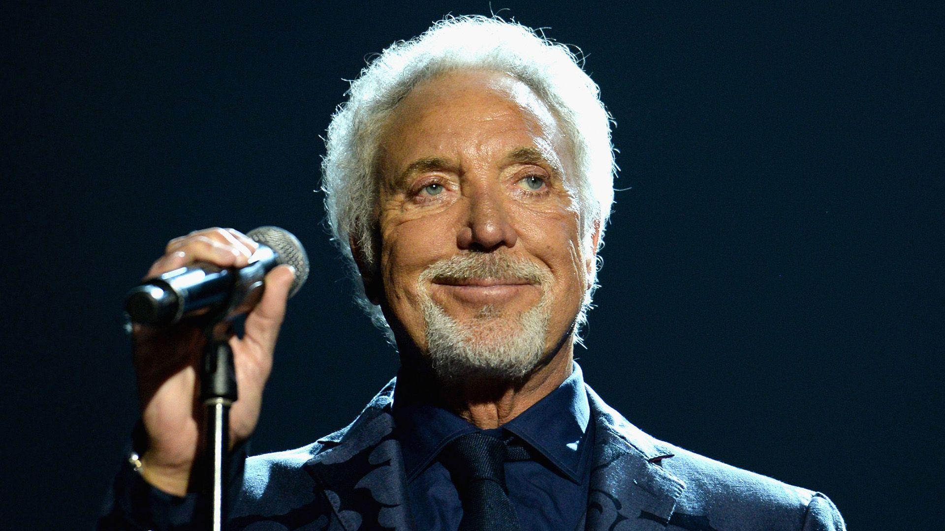 Sir Tom Jones holds a microphone and smiles at the audience. He is wearing a dark blue suit, shirt and tie, and looking away from the camera.