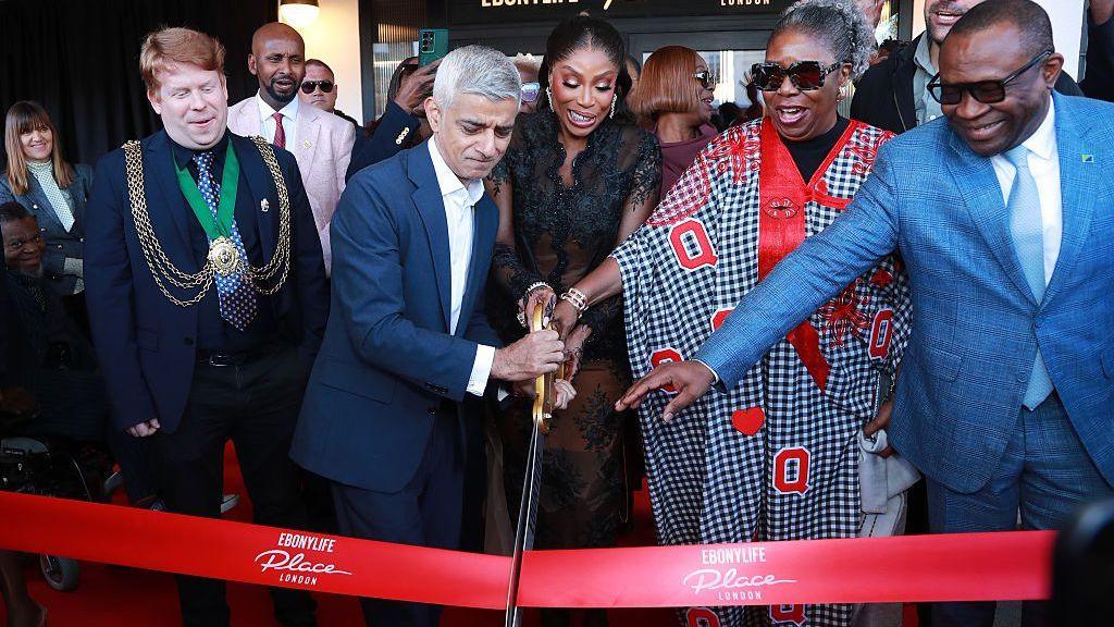 Sadiq Khan and Mo Abudu cut a red ribbon with giant golden scissors, flanked by guests during the opening ceremony for EbonyLife Place London.