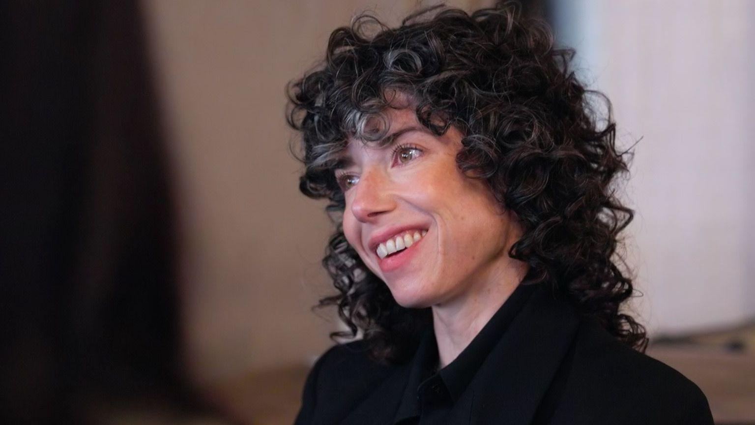 Meredith Whittaker smiling and looking to one side, with dark, curly hair and wearing a black shirt.