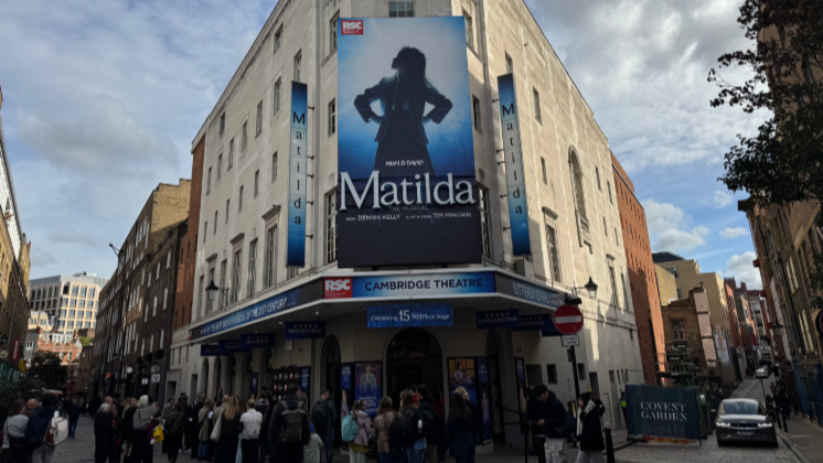 A blue and black advertising hoard for the musical stands outside and above the entrance area of a London theatre. Crowds mingle below. It is daytime.