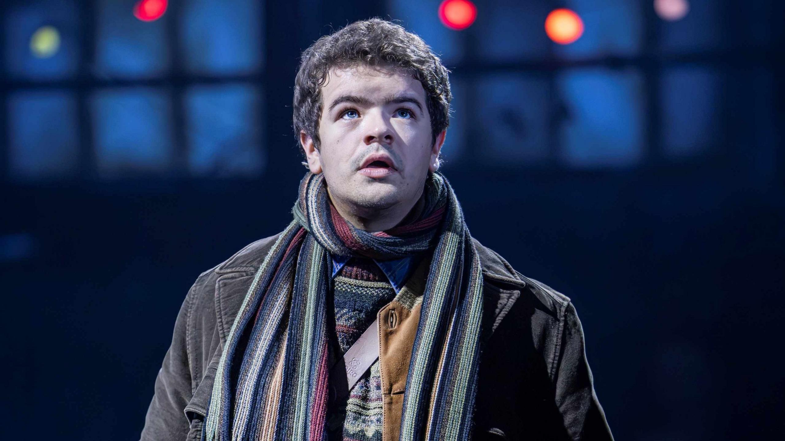 Gaten Matarazzo on stage as Mark in Rent. He wears a colourful striped scarf, patterned jumper, and brown jacket.