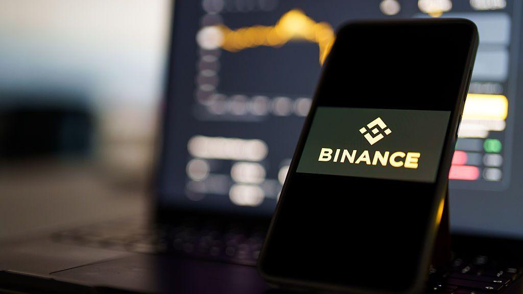 A phone propped up in front of a laptop. On the phone screen it says Binance and the blurred laptop screen shows a bar chart