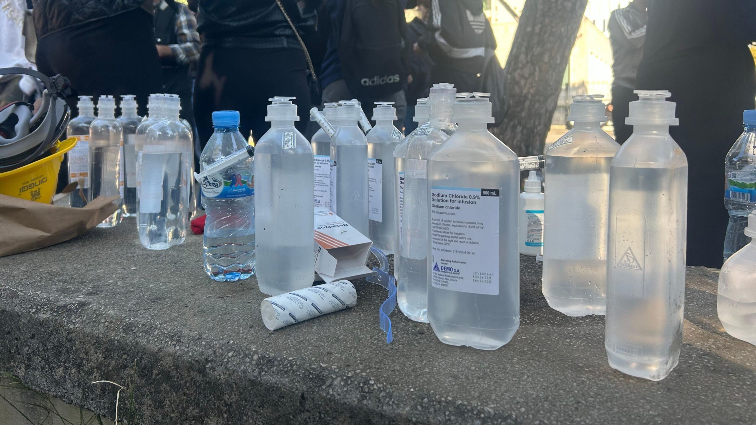Clear bottles of saline solution are arrayed over a flat stone surface, with a group of people standing in sunlight nearby.