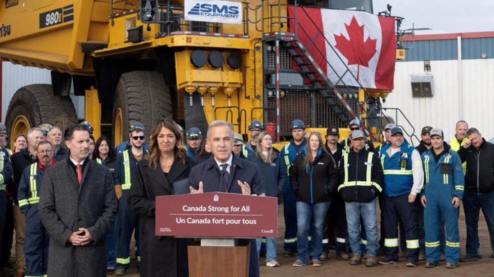 Mark Carney stands before a podium with a sign that reads in French and English: 'Canada Strong for All'.  He is flanked by Alberta Premier Danielle Smith, and behind him are oil workers in hard hats and work vests. A large piece of yellow equipment is hung with a Canadian flag.