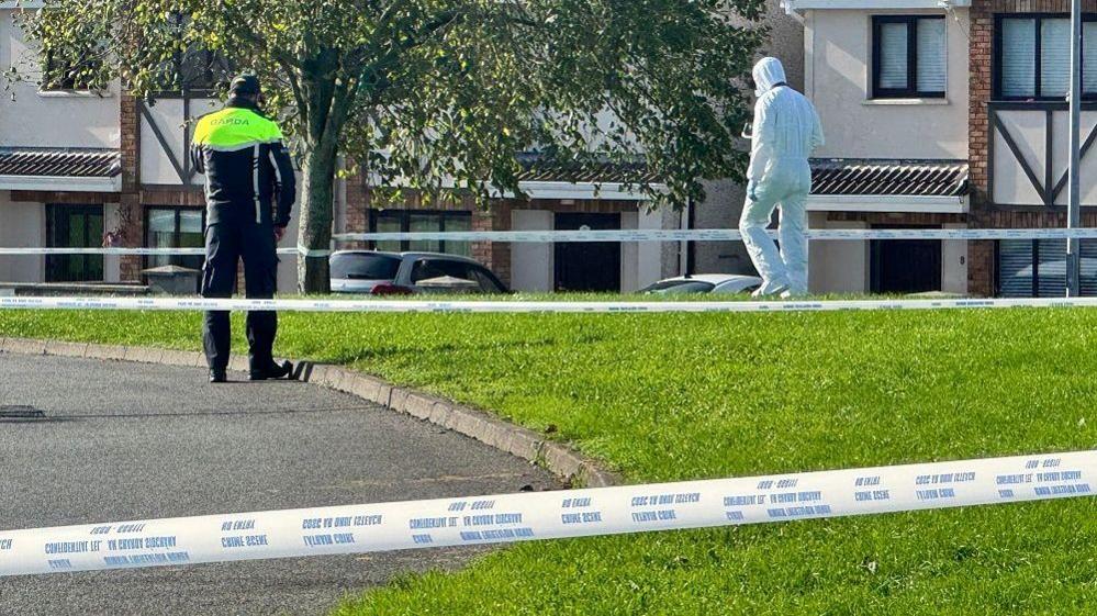 A man wearing a navy and high-viz yellow GARDA jacket, with navy trousers, stood in a housing estate. There is also a man in a full white forensics outfit, and multiple lengths of police tape cordoning off the area.