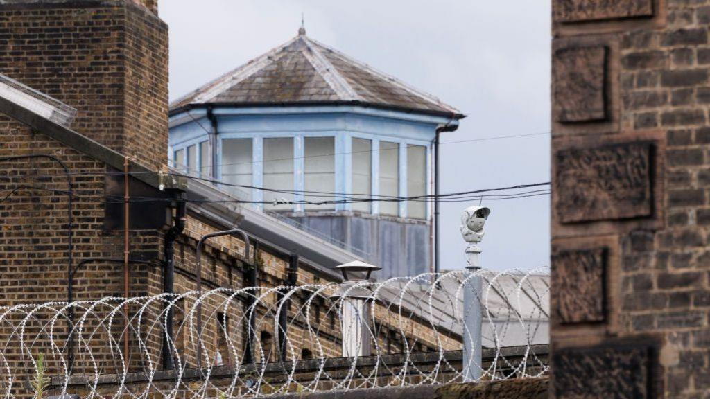 Barbed-wired fencing above brown brick walls with a security cameras are pictured at the HMP Wandsworth prison in London, England.