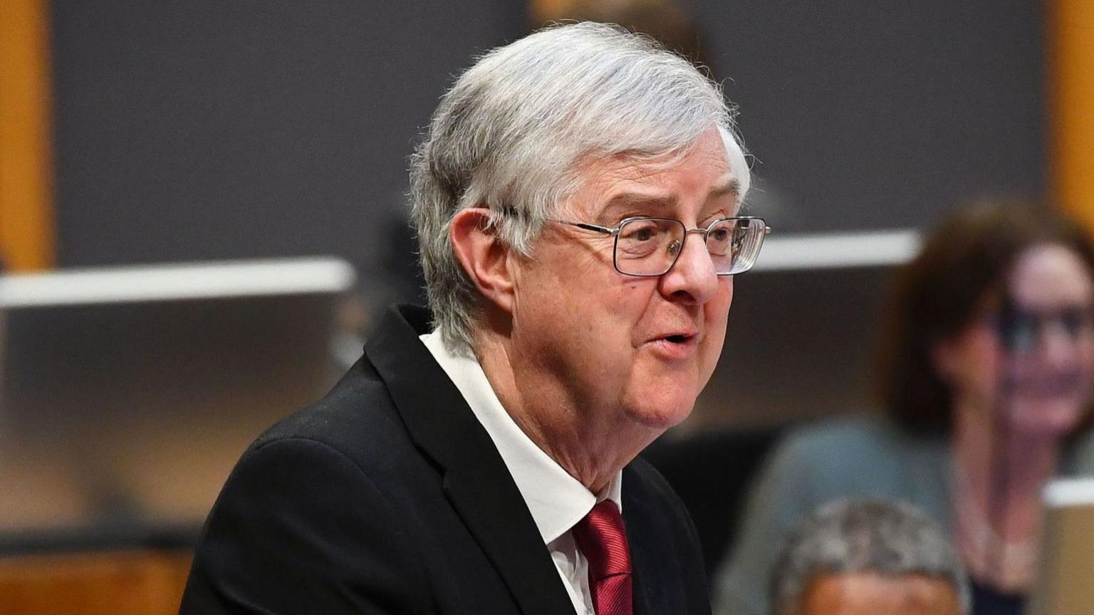 Mark Drakeford speaking in the Senedd chamber. He is shown from the chest up, turned slightly to the right of the camera. He has short grey hair, is wearing rectangular glasses and a dark suit jacket, white shirt, and red tie.