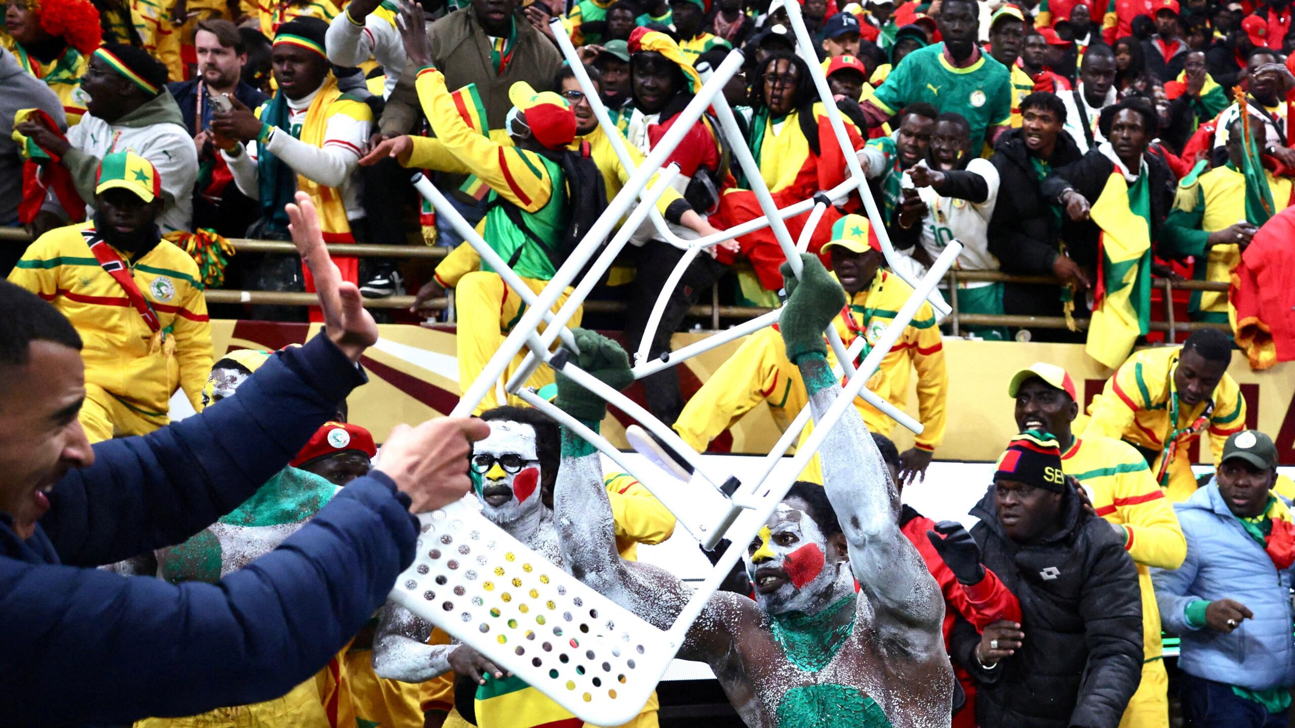 Senegal supporters clash with security personnel as they attempt to storm the field of play after a penalty decision against their team during the 2025 Africa Cup of Nations final in Rabat. In the central foreground is a man covered in white body paint with green, yellow and red dots launching a white chair at a man in a blue puffer jacket who is grimacing, holding up his left hand in an effort to protect himself while also wielding a similar chair with his right hand. Behind the two central characters are several other Senegal supporters, some wearing all-yellow tracksuits and baseball caps, who have come out of the stand and are coming over hoardings to reach the ground. In the background is the packed stand full of Senegal supporters, many of whom are gesticulating in an animated way