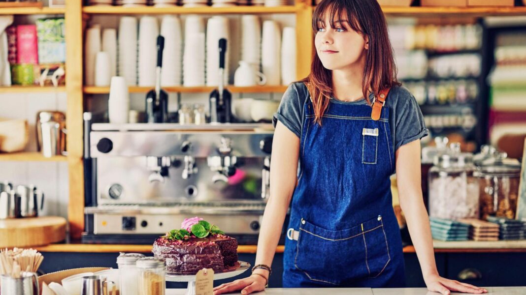 How much is the minimum wage going up by? A young woman with long reddish-brown hair and wearing a dark blue apron stands behind the counter in a brightly-lit cafe, next to an ornate chocolate cake. There is a large coffee machine on the back wall, with piles of empty take-away cups and several boxes of tea bags.