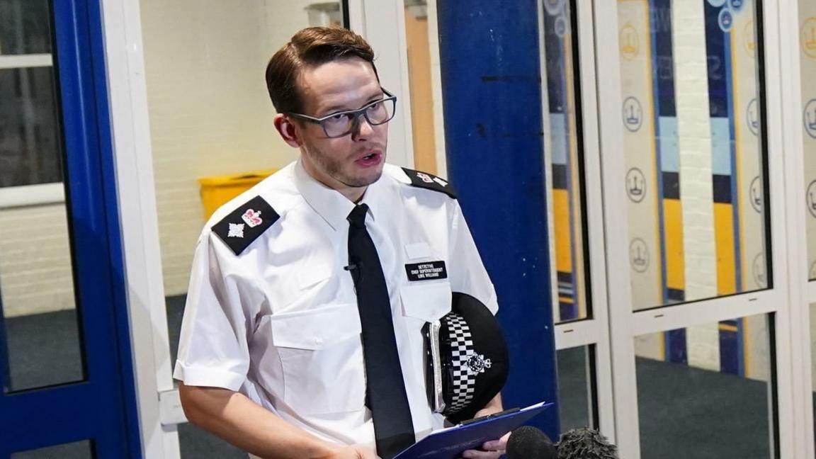 Luke Williams wearing a white short-sleeved police uniform shirt and tie, holding a peaked cap under his arm while speaking to reporters.