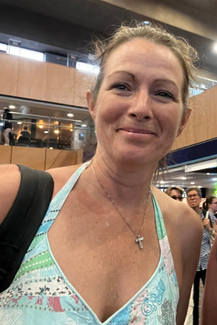 A selfie shot of a woman smiling at the camera. She has a blue strap top on and a black strap for a backpack on the left shoulder. She has a necklace on with a cross pendant. It is a head and shoulders shot.