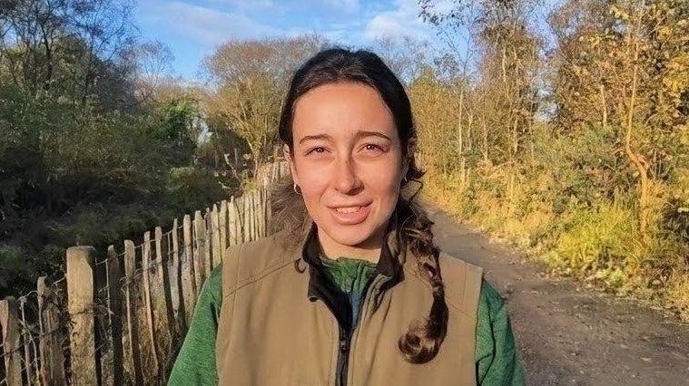 Chloe McElhatton has long brown hair and is smiling. She is wearing a tan gillet and green shirt. She is standing on a path with a wooden fence and large trees.