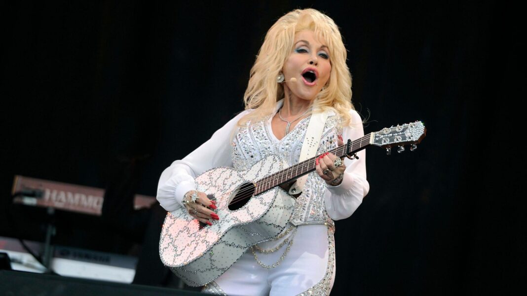 Dolly Parton is holding an acoustic guitar and wearing a white outfit while singing on stage.