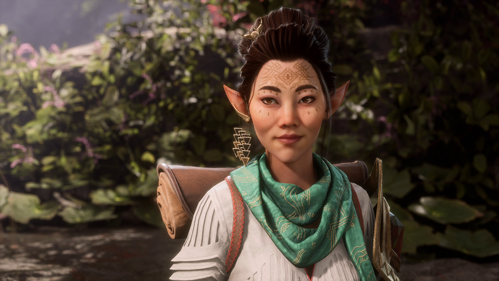 A character from Dragon Age with pointy, elven ears and a henna-type tattoo on their forehead stands in a woody clearing in a forest. They appear to be on their way somewhere, carrying a leathery roll-top backpack and wearing a green neckerchief.