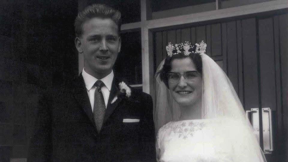 A black-and-white wedding photograph showing two people standing side by side outside a building with panelled doors and large window frames. Rex, on the left, is wearing a dark suit, light-coloured shirt, tie and a buttonhole flower. Kay, on the right, is wearing a wedding dress with a veil, glasses and a floral headpiece. Both are facing the camera,