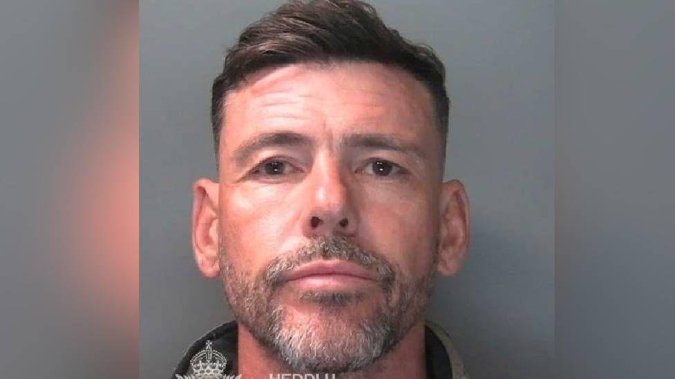 A police mugshot of Gavin Smith. The man in the a head-and-shoulders image has short brown hair, a greying, stubbly beard and a neutral expression.