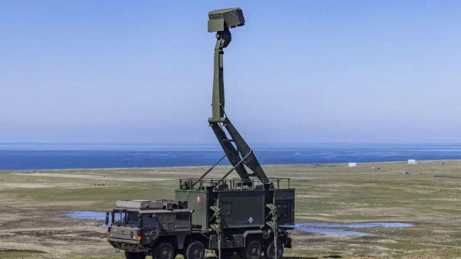 A green military vehicle sits in a field - on top of the vehicle is a tall green defence system that reaches into the sky. The ocean is visible in the distance.