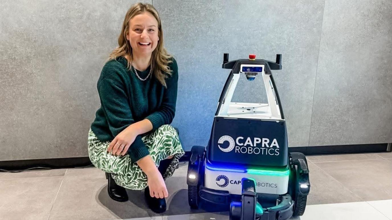 Lauren Townsend posing beside a waist-height robot, which is coloured white and navy. Lauren wears a green jumper and floral green skirt. She has blonde hair past her shoulders.