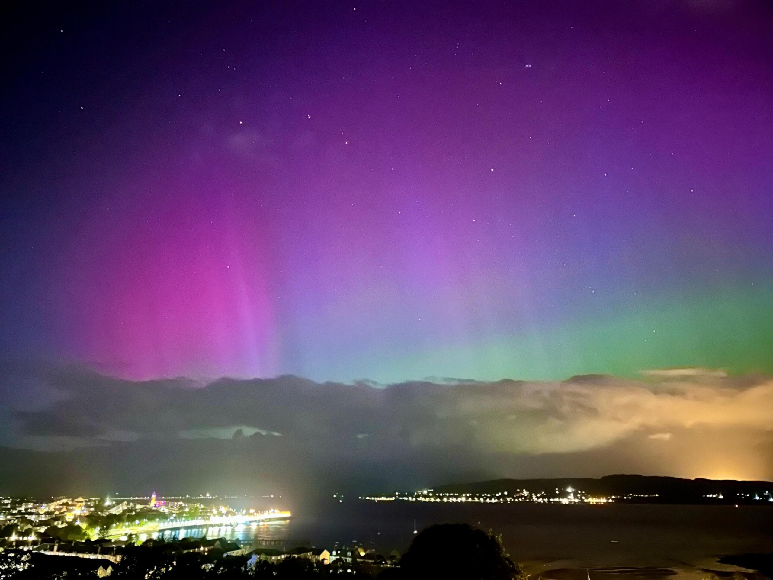An image of the sky in Gourock, Inverclyde, on 10 October during the evening. The Northern Lights can be seen in a variety of pink, blue and green hazes above clouds and the city.